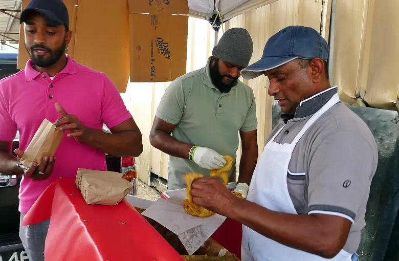 A vendor wrapping doubles in paper at a roadside stall in Trinidad while two customers wait.