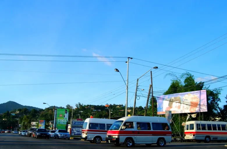 A line of maxi taxis waiting at a Trinidad terminus, their route color bands along the side, with hills behind.