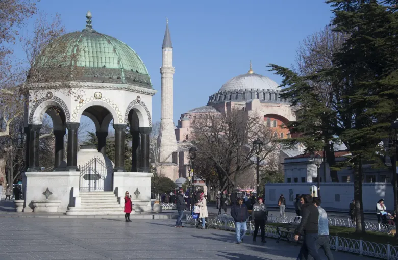 People walking the flat paved square at Sultanahmet in Istanbul, Turkey, past the German Fountain with Hagia Sophia behind.