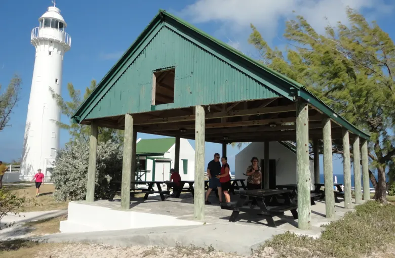 The white Grand Turk Lighthouse beside a green roofed shelter with picnic tables, where visitors sit in the shade.