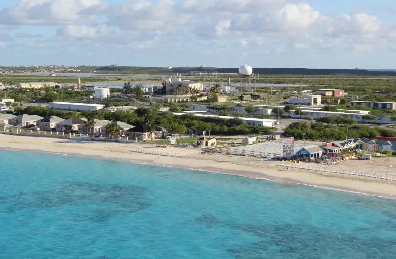 Low buildings along the shore of Grand Turk above a beach and clear turquoise water, seen from offshore.