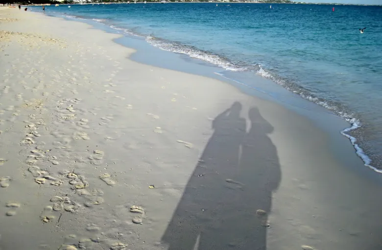 The long shadows of two people cast across rippled pale sand at the water's edge on a bright afternoon.