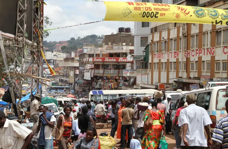 Crowds, stalls and minibuses filling a busy street market beneath shopfronts in Uganda.