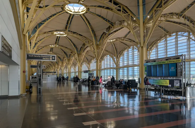 A vaulted airport hall with a South Security Checkpoint sign, departure boards and travelers walking with bags.