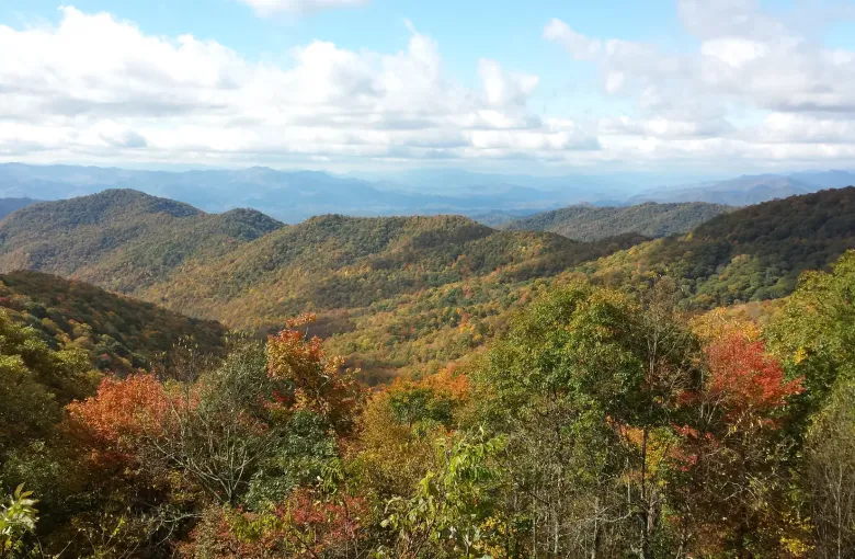 Forested ridges in orange, red and green fall color receding into blue haze under a partly clouded sky.