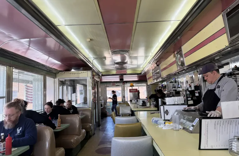 The narrow interior of a railcar diner, with a cook at the grill behind a long counter and customers eating in booths.