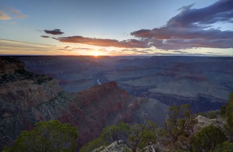The sun setting between layered canyon walls, with the Colorado River visible on the floor far below.