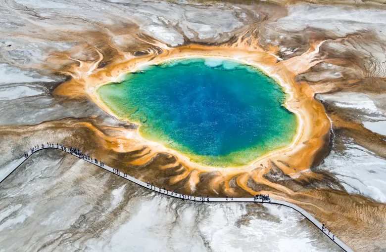 An aerial view of a wide hot spring ringed in orange and gold, with visitors walking the boardwalk that curves past it.