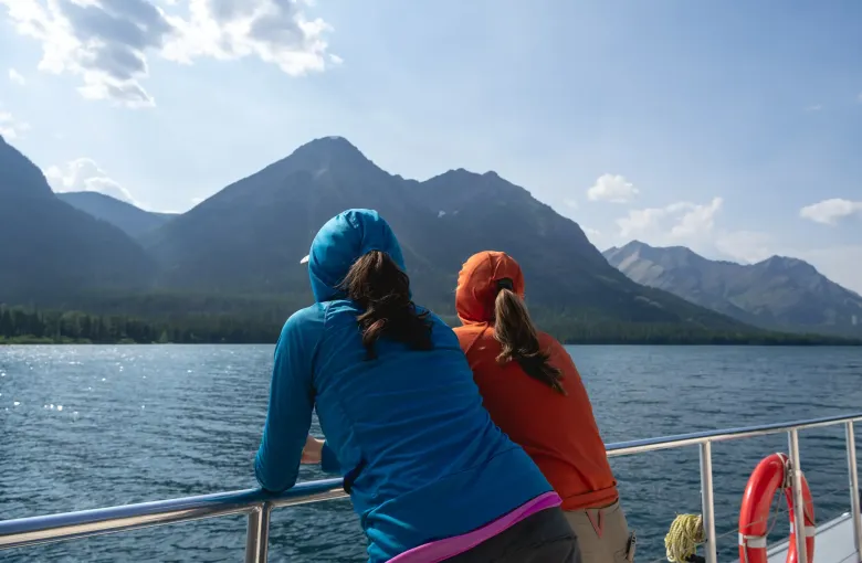 Two people in hooded fleece layers lean on a boat rail, looking across a lake toward snow streaked mountains.