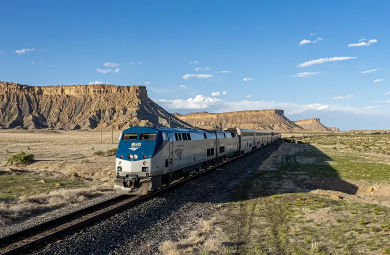 A silver and blue passenger train on a single track crossing open scrubland beneath a flat topped desert escarpment.