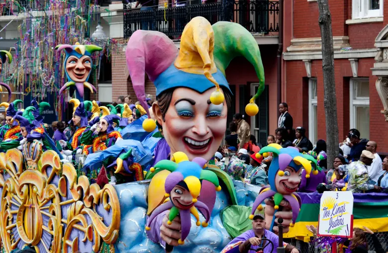 A carnival float carrying giant jester heads in purple, green and gold passes a crowd on a New Orleans street.