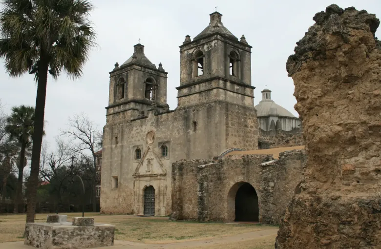 A weathered stone mission church with twin bell towers and a dome, seen past a ruined wall and palm trees.