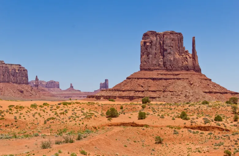 A tall red sandstone butte rising from an orange desert floor, with smaller buttes on the horizon under a clear sky.