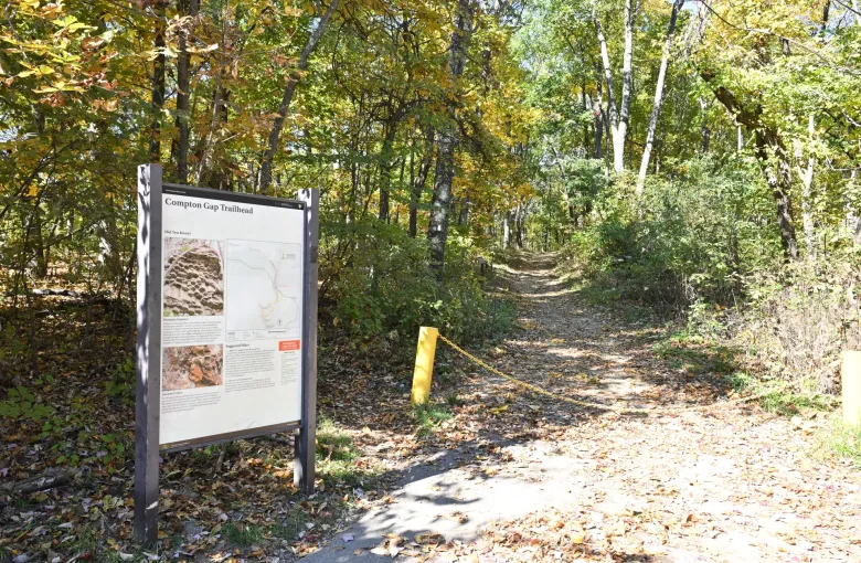 A trailhead board carrying a route map, suggested hikes and a red emergency telephone panel beside a leaf covered path.