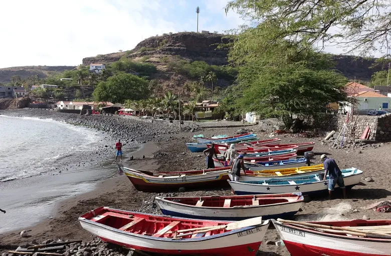 Painted open boats drawn up on the dark beach at Cidade Velha, with the fortified ridge above.