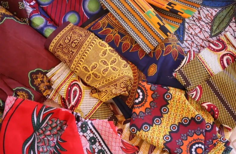 Lengths of printed capulana cloth stacked in overlapping folds, in deep reds, yellows, blues and greens.