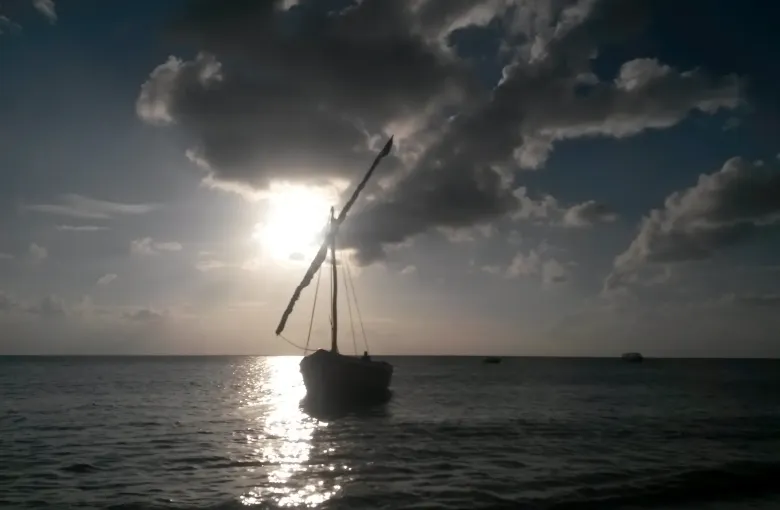 A dhow lying at anchor in silhouette, the sun breaking through heavy cloud and laying a track of light across the water.