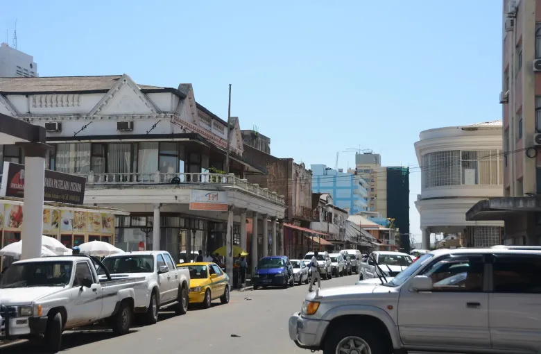 Colonnaded shopfronts along a downtown Maputo street, with taxis and pickups parked along both curbs.