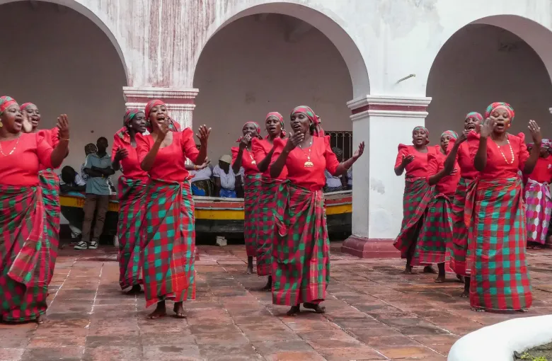 Women in matching red tops and checked capulana wraps singing and clapping in an arcaded courtyard in Mozambique.
