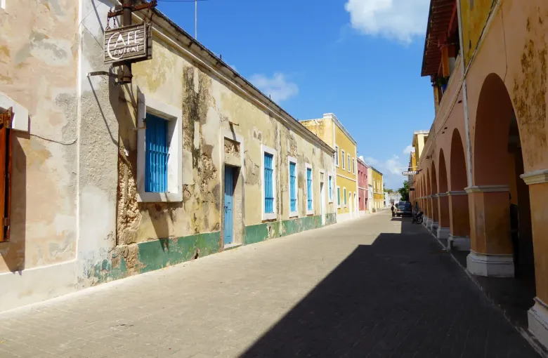 Weathered lime-washed facades with blue shutters along a paved street on the Island of Mozambique, an arcade opposite.