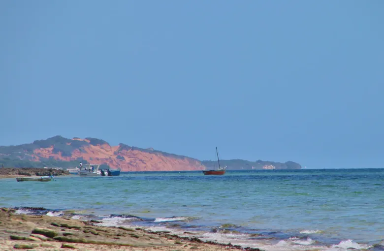 Red dune cliffs across the water at Vilankulo, with a dhow and small boats anchored in the shallows under a clear sky.