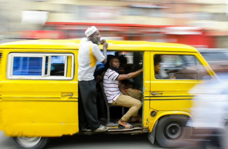 A yellow danfo minibus in motion on a Lagos street with its conductor standing in the open doorway.